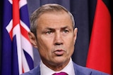 A tight head shot of Roger Cook with a serious expression at a media conference