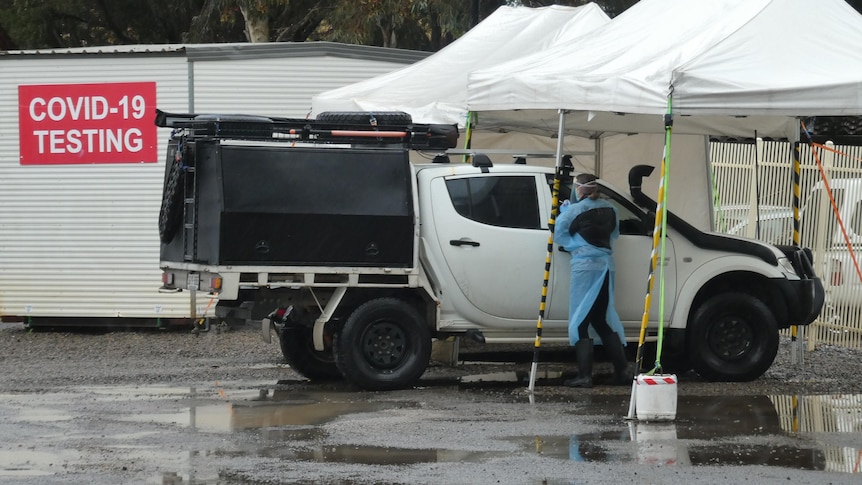 A person in PPE stands next to a white ute in front of a temporary building with a COVID TESTING sign