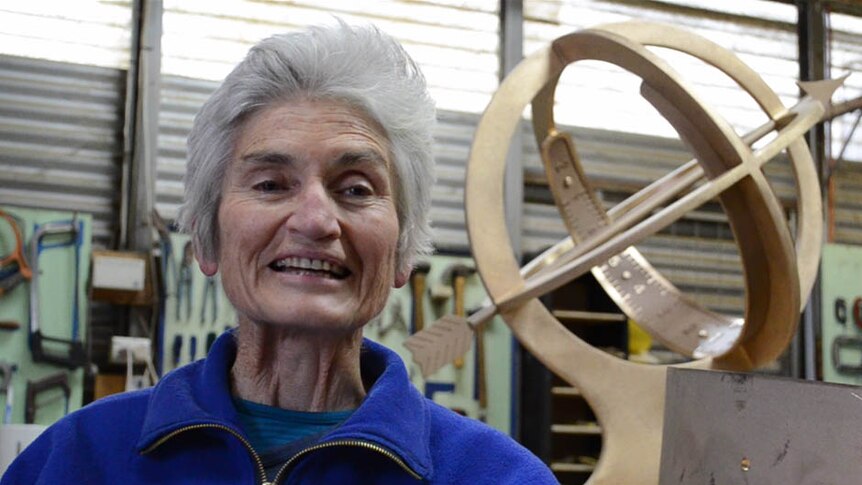 A smiling woman with white whispy hair sits in a shed, with tools on the wall and a large brass spherical sundial behind her.