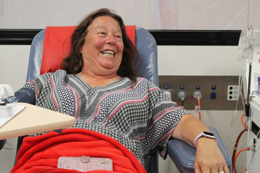 A woman sits in a blood donation chair wearing a patterned top looking smiley to the right of the camera