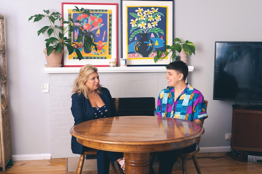 A woman wearing a dark blazer sits across a table from a young person wearing a bright shirt