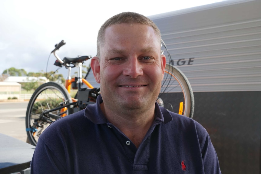 A man faces the camera standing in front of a caravan and bikes.
