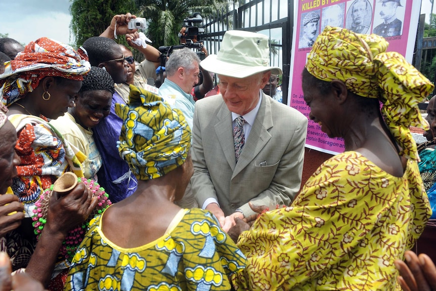 A man shakes hands with a number of smiling Nigerian women. They are wearing colourful traditional clothes, he is wearing a suit