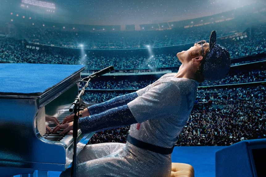 The actor, in a sequined baseball uniform, sitting at a piano, looks out to a huge stadium sized audience.