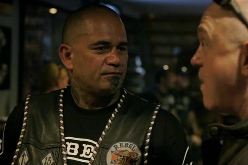 A man in a Rebels vest speaks to another man inside a motorcycle clubhouse.