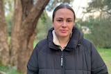A woman with brown hair wearing a black puffer jack looks at the camera with trees in the background