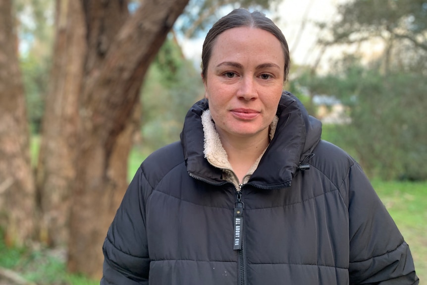 A woman with brown hair wearing a black puffer jack looks at the camera with trees in the background