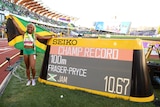A smiling Jamaican sprinter holds a flag behind her standing next to a scoreboard saying "Champ record 100m Fraser-Pryce JAM".