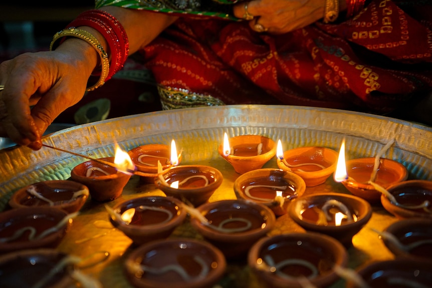 A hand holding a lighted match to spark a flame over a clay oil lamp, sitting in a tray with more clay oil lamps