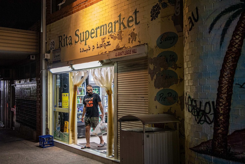 A man carrying a shopping bag walks out of a yellow brick shop with 'Rita Supermarket' written above the door.