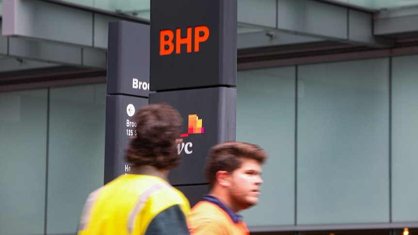 Two men in high-vis walk past a BHP sign in a city.