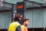 Two men in high-vis walk past a BHP sign in a city.