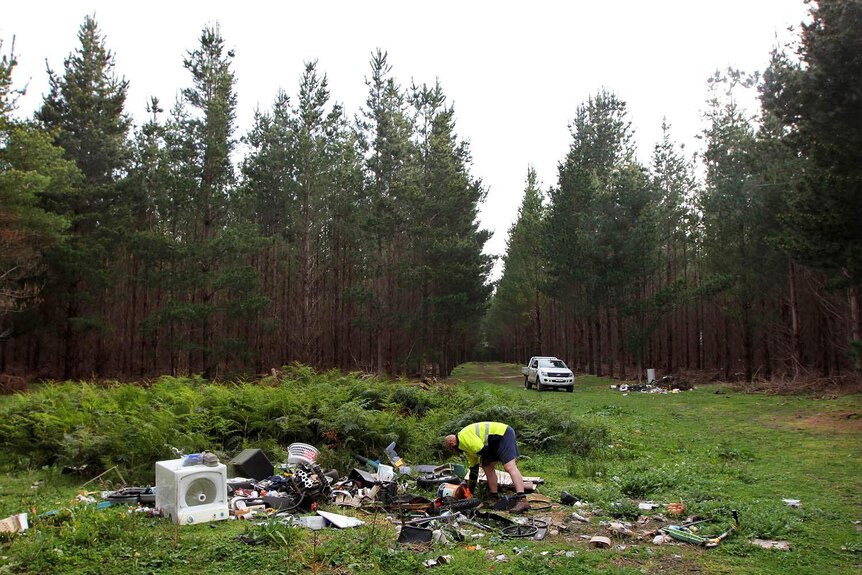 A man in yellow high-visibility gear bends over a large pile of rubbish in a forest, with more piles visible in background.