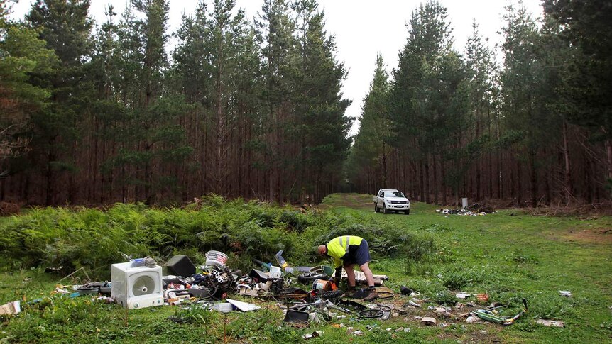 A man in yellow high-visibility gear bends over a large pile of rubbish in a forest, with more piles visible in background.