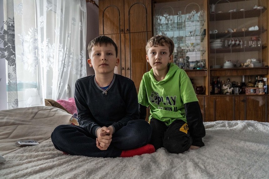 Two little boys sit on a bed, one cross-legged and one kneeling, looking at the camera with serious expressions