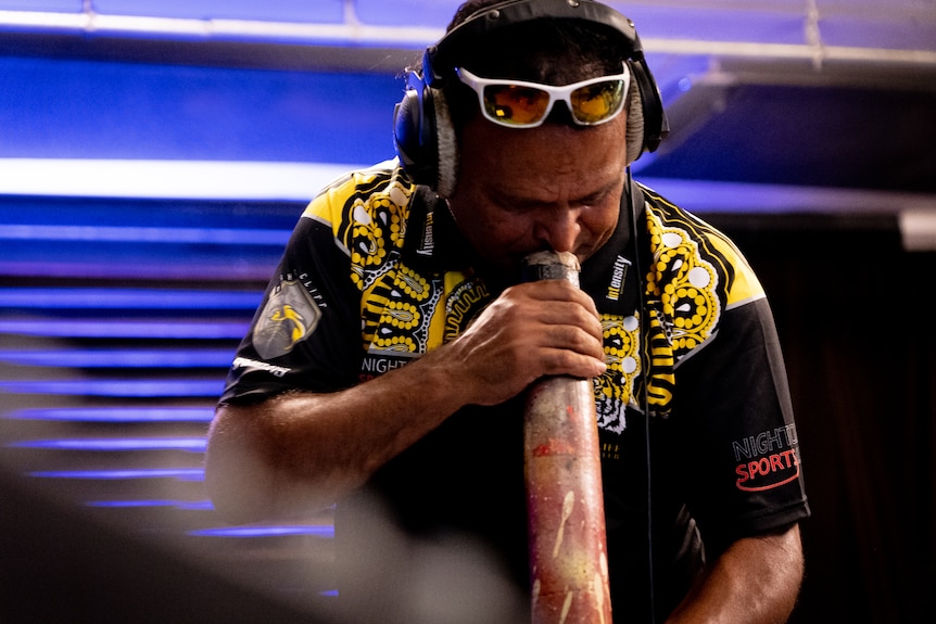 A man with short dark hair leans over playing a didgeridoo 