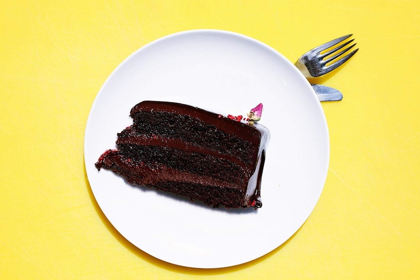 A shiny red/brown piece of chocolate cake on a white plate, on top of a bright yellow table.