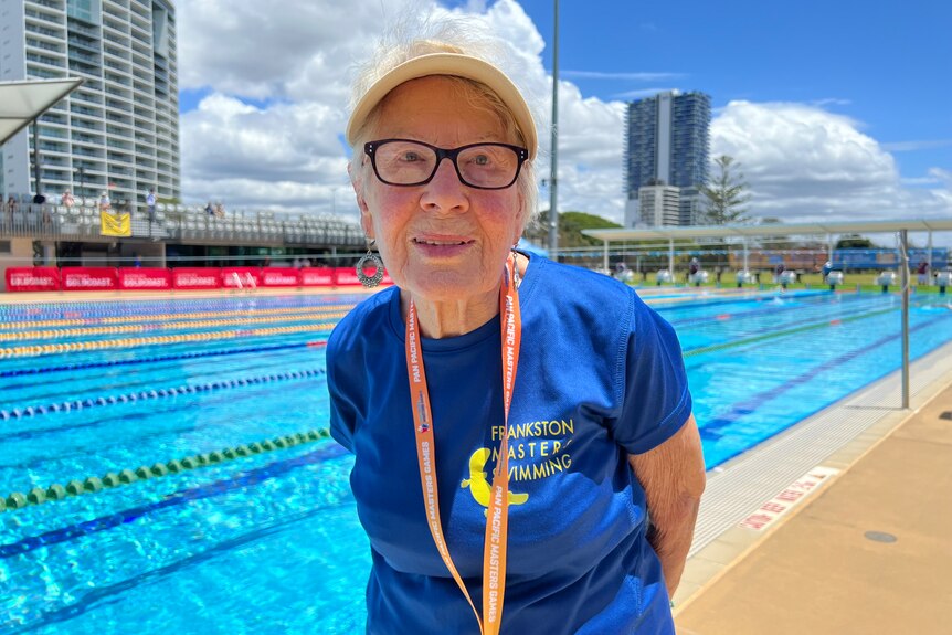 An elderly woman with black glasses wearing a yellow visor, standing in front of a blue pool