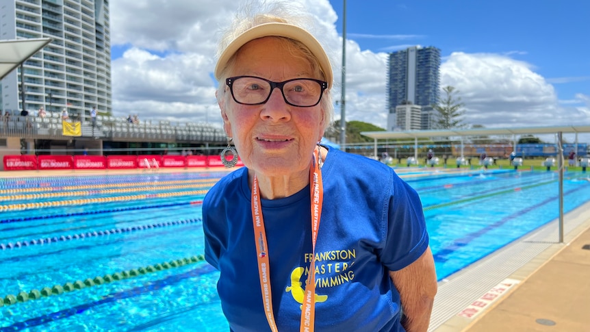An elderly woman with black glasses wearing a yellow visor, standing in front of a blue pool