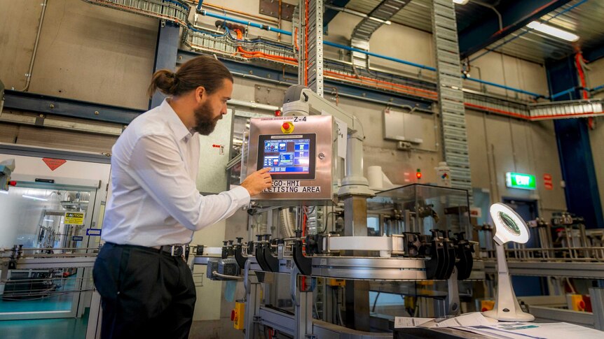 A man in business attire and with a ponytail presses a button on a screen. Around him is an assortment of machinery.