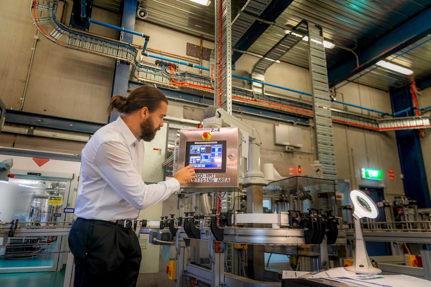 A man in business attire and with a ponytail presses a button on a screen. Around him is an assortment of machinery.