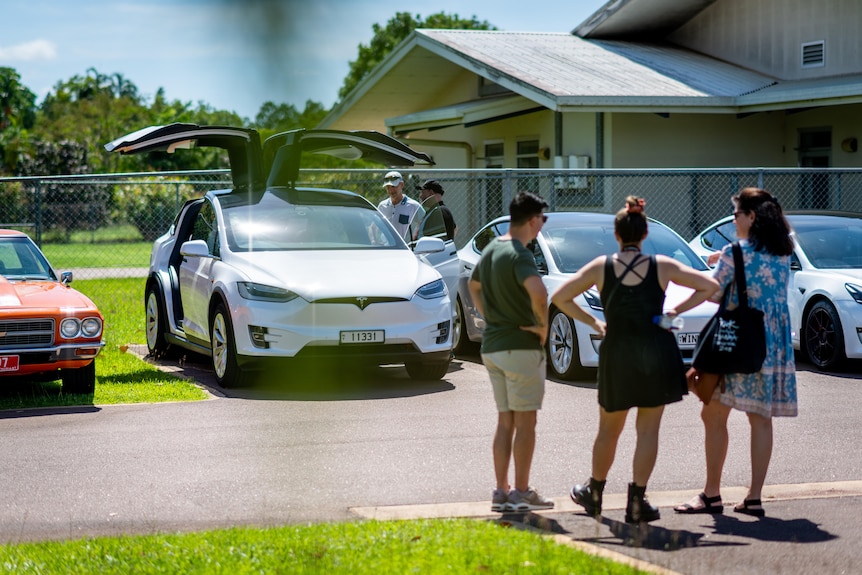 Three people are standing in a parking lot near a Tesla vehicle with the doors open.