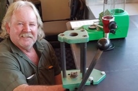 a man with fair long hair crouches next to a workbench and a vice