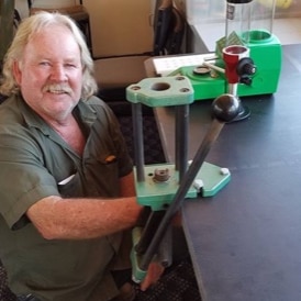 a man with fair long hair crouches next to a workbench and a vice