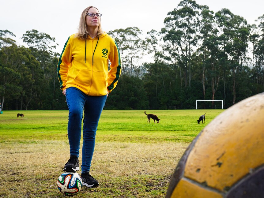 A woman wearing a yellow Matildas jacket has her foot on a soccer ball, she looks into the distance. 