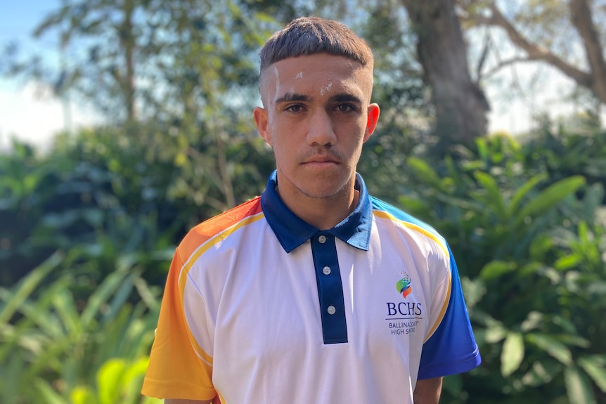 An Indigenous teenage boy with short dark hair wearing a white blue and yellow school shirt.