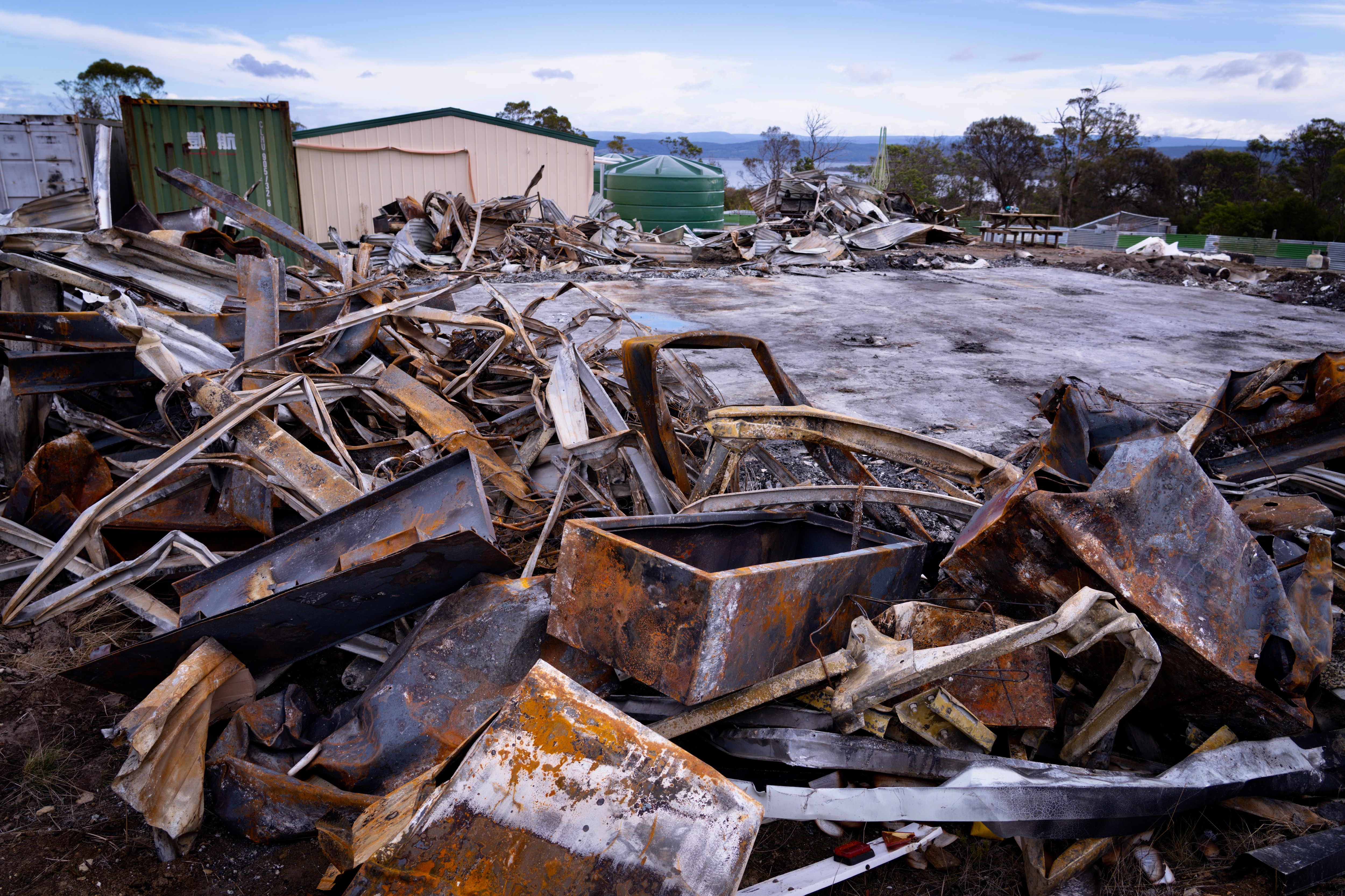 The burnt rubble of a house. In the background, a shed and shipping container still stand.