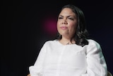 A photo showing an Indigenous female senator wearing a white dress with a dark background