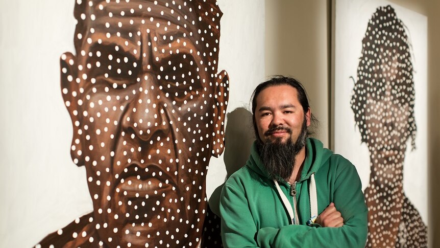 Indigenous artist Damien Shen standing in front of two paintings hanging on a wall