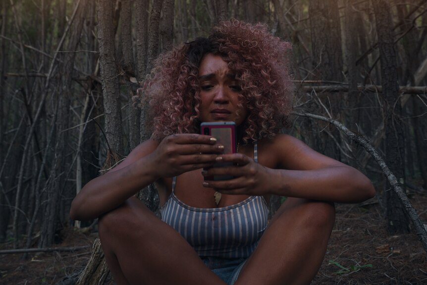 Young woman of colour with dyed pink curls sits holding phone with a look of despair in thick woods.