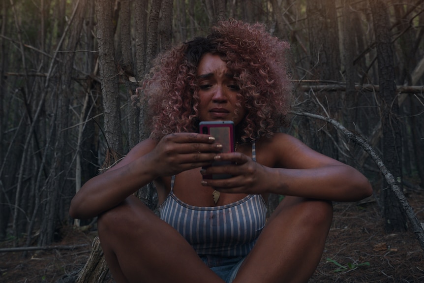 Young woman of colour with dyed pink curls sits holding phone with a look of despair in thick woods.