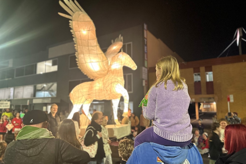 A young girl sits on the shoulders of a man as a large lantern of a flying horse passes in front of her