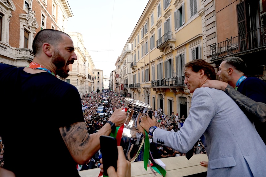 Un joueur de football italien et l'entraîneur de l'équipe nationale sur un balcon tentent de soulever le trophée de l'Euro 2020 avec la foule en dessous.