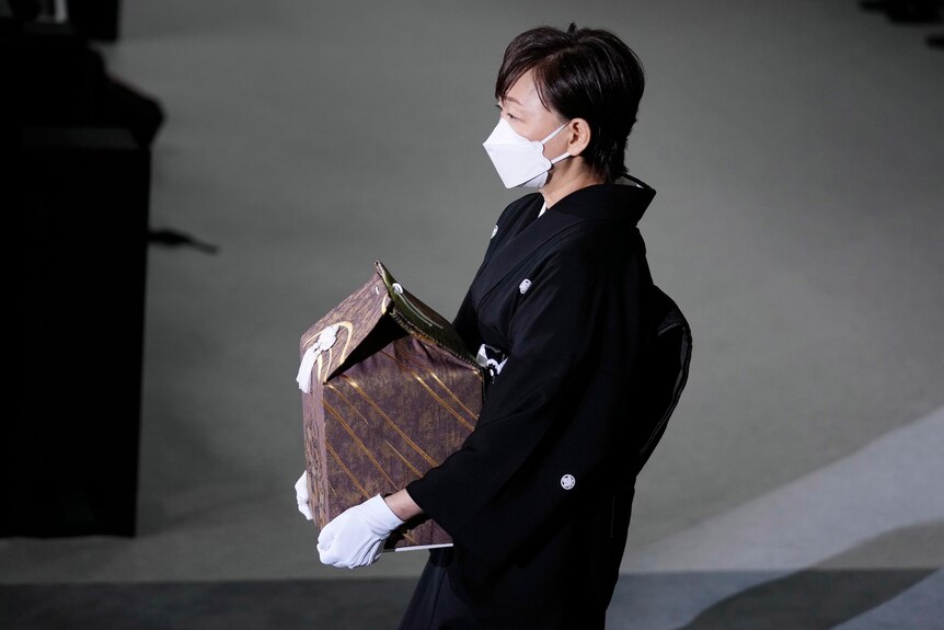 A woman in dark clothes, white gloves and face mask hold a ceremonial box as she walks.