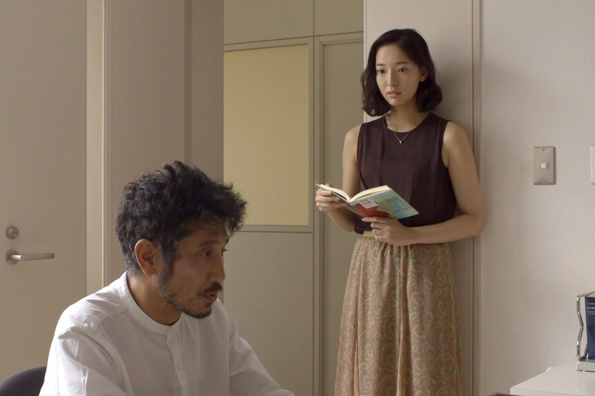 Japanese woman in dark purple singlet and long beige skirt holds book and looks apprehensively at Japanese man in white shirt.