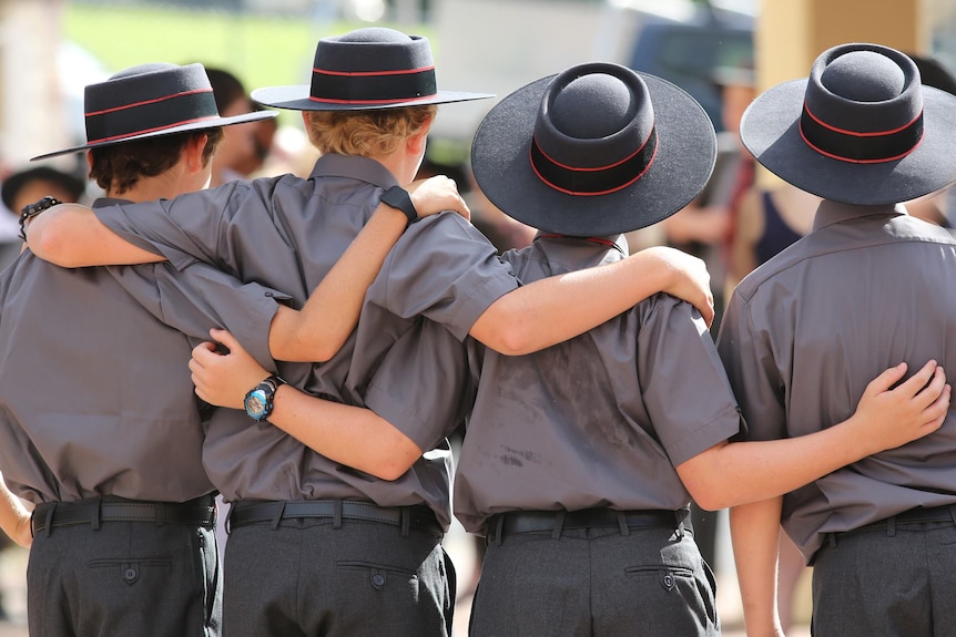 Four boys in school uniforms - a brim hat, shorts and collared shirt - stand with backs turned and arms around each other.