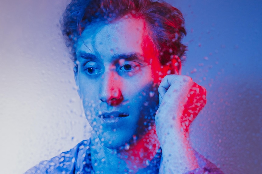 A man bathed in blue light sits in front of a piece of glass covered in raindrops