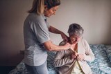 An aged care working standing up helps dress a senior woman sitting on her bed.