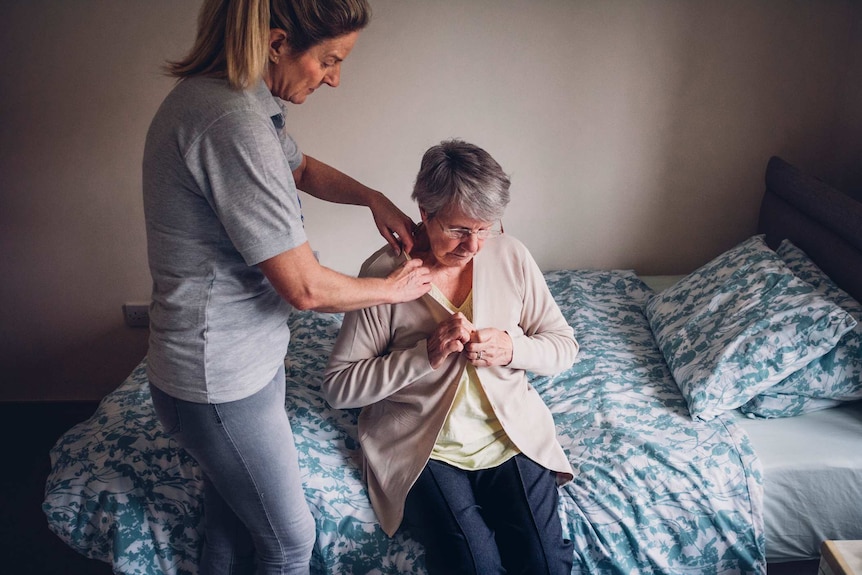 An aged care working standing up helps dress a senior woman sitting on her bed.