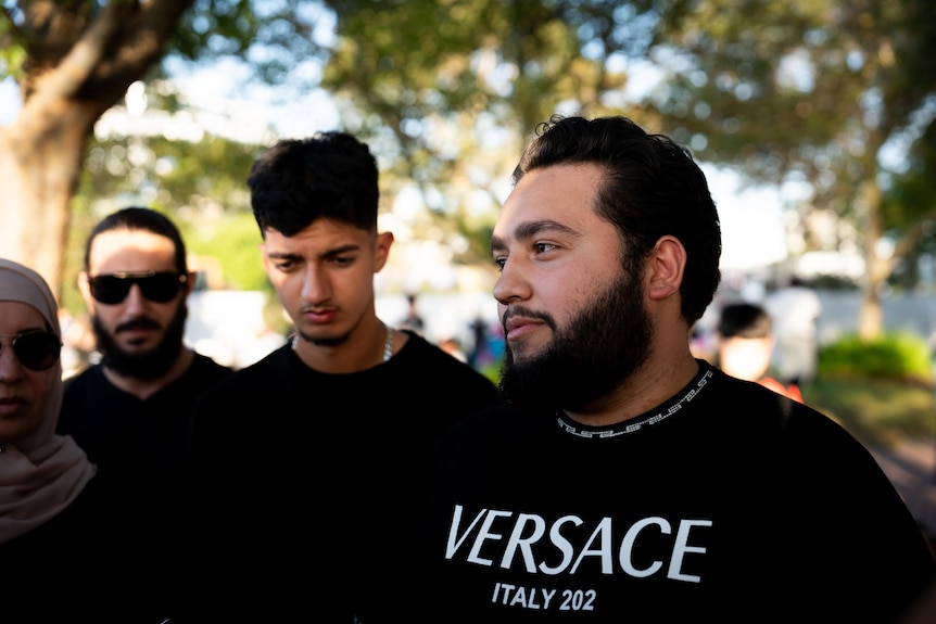 A young man with a beard in a black Versace T-shirt stands in a park.