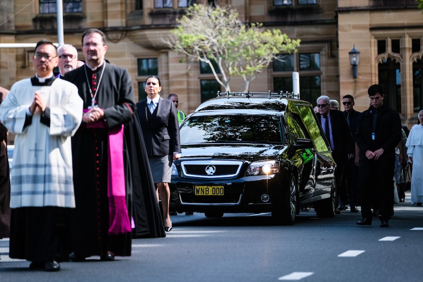 Clergymen walk in front of a black hearse driven down a quiet road