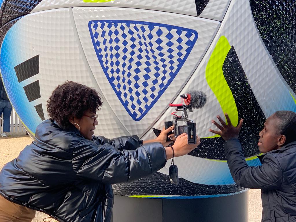 Woman holding a mobile phone with a microphone attached filming a woman putting her hand on a picture of a giant soccer ball.