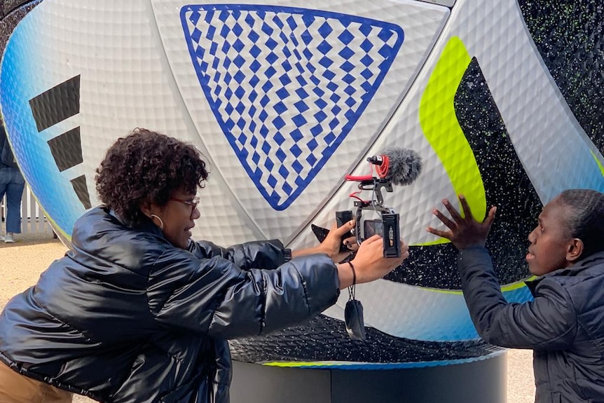 Woman holding a mobile phone with a microphone attached filming a woman putting her hand on a picture of a giant soccer ball.