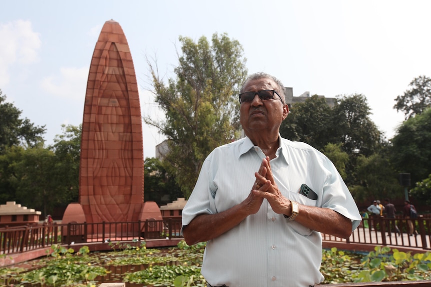 A man with his hands bowed and wearing sunglasses looks into the distance in front of a surfboard shaped structure.