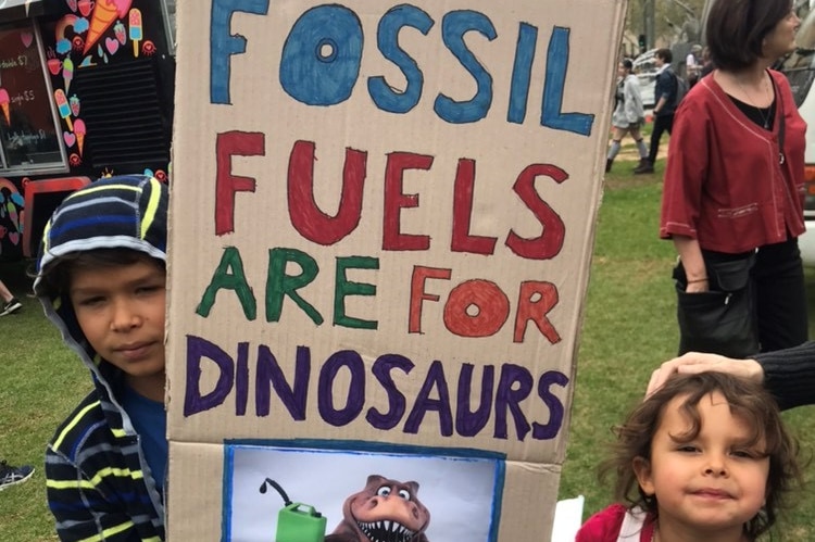 A young boy in a colourful hoodie holds a sign that says 'fossil fuels are for dinosaurs'.