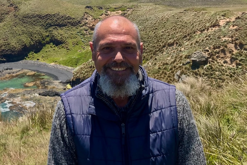 A man with a beard and grey hear smiling and standing at the top of a hill, with a lush green landscape with an ocean
